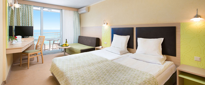 Durch das helle Zimmer führt der Blick über den Balkon auf den Strand und das dahinterliegende Meer, das ruhig die Küstenlinie von Albena begleitet.