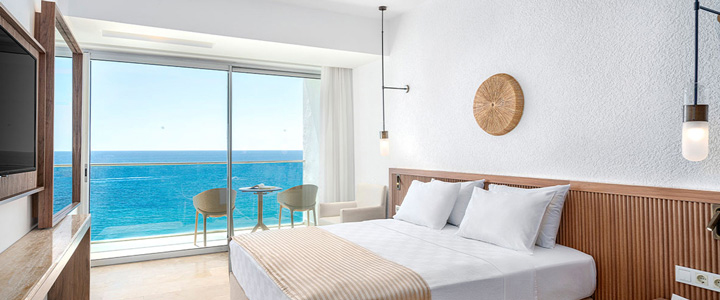 Die moderne Suite mit klaren weißen Linien und Holzelementen öffnet sich über eine große Glasfront zum Balkon, mit ruhigem Blick auf das Mittelmeer.